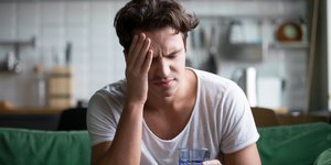Νεαρός άνδρας με έντονο πονοκέφαλο κάθεται με ένα ποτήρι νερό στην κουζίνα,