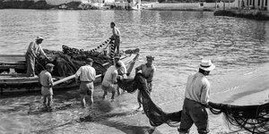 Μύκονος, 1955. Ξημερώματα στον μόλο Καμπάνη, ψαράδες ξεφορτώνουν τα δίχτυα τους για στέγνωμα και επισκευή. Το νεόκτιστο ξενοδοχείο Λητώ διαφαίνεται στο βάθος