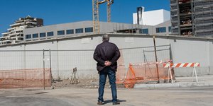 Ένας άνδρας «umarell» στέκεται με τα χέρια πίσω από την πλάτη, κοιτάζοντας πίσω από φράκτη έργων