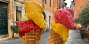 Δύο άτομα κρατούν πολύχρωμα παγωτά gelato σε ένα λιθόστρωτο δρόμο στη Ρώμη, με ιστορικά κτίρια στο παρασκήνιο